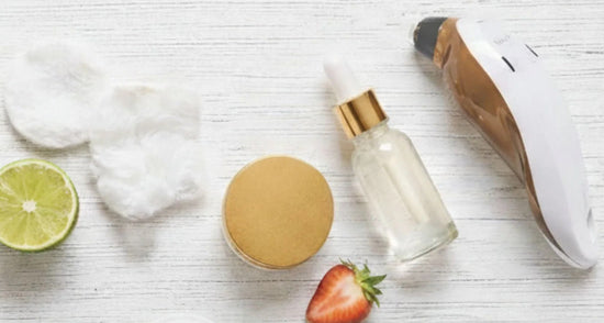 Skincare products, serum bottle, cotton pads, facial device, lime and strawberry on white wood.
